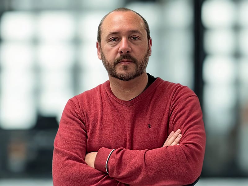Un hombre con barba y jersey rojo permanece de pie con los brazos cruzados, mirando a la cámara en el interior, dispuesto a dirigir un taller de inteligencia artificial.
