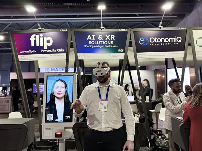 Un hombre utiliza un casco de realidad virtual en un stand de una exposición tecnológica, explorando pantallas digitales impulsadas por inteligencia artificial, con gente de fondo.