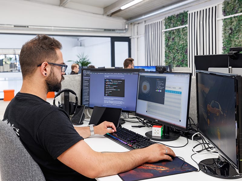 Hombre con gafas trabaja en un escritorio durante un taller de inteligencia artificial, rodeado de tres monitores que muestran código y gráficos en una oficina moderna.