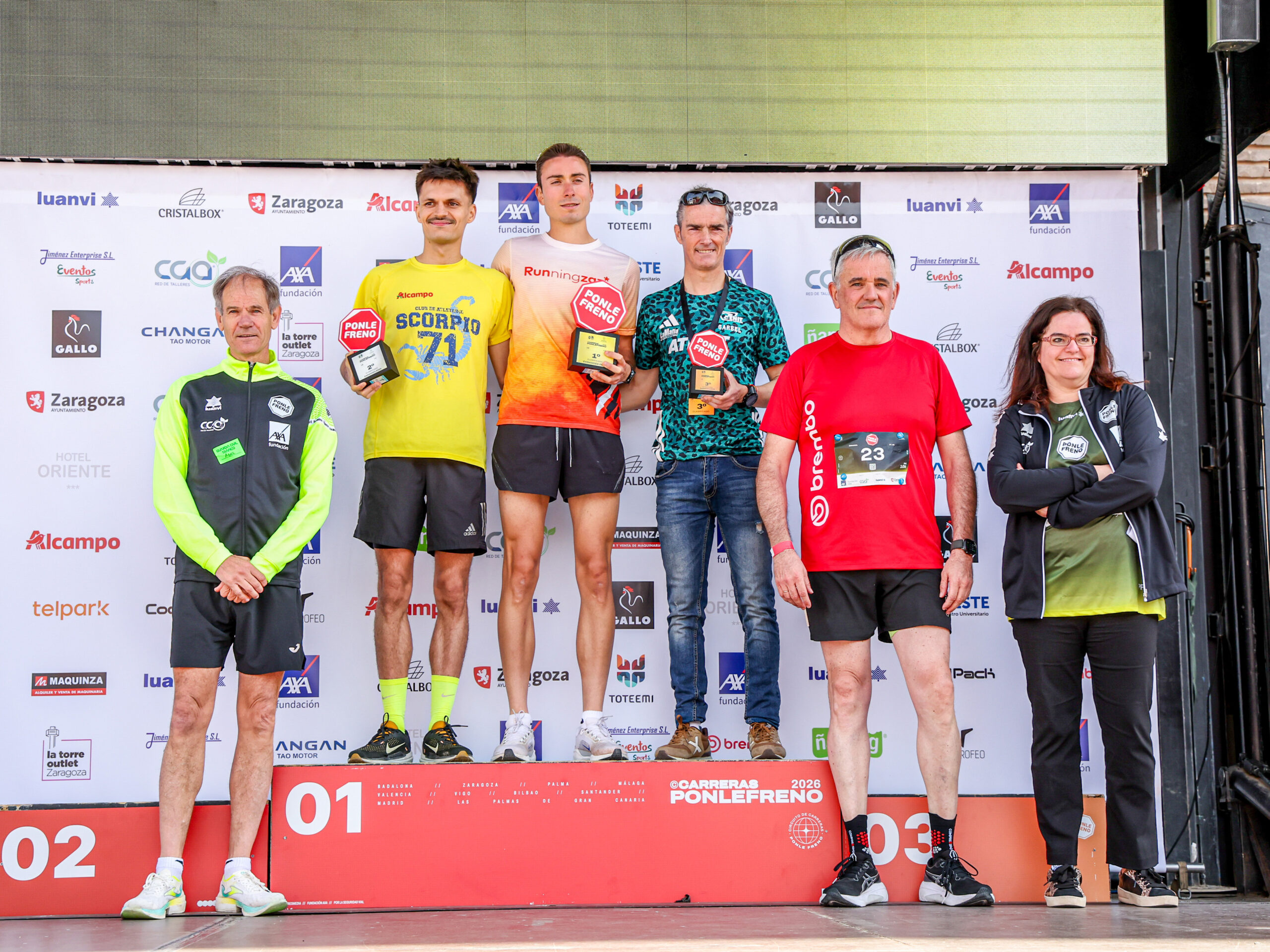 Seis personas de pie en un escenario en una carrera, con tres hombres sosteniendo trofeos en un podio.