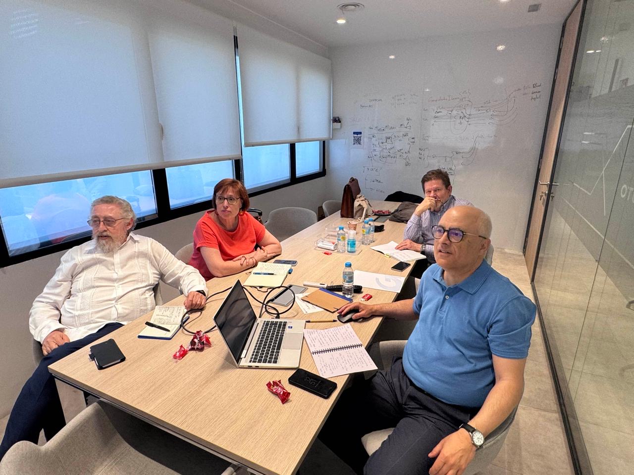 Cuatro personas se sientan alrededor de una mesa de reuniones con ordenadores portátiles, papeles y bebidas, mirando hacia la cámara mientras discuten los planes de EuroPremium 2026.