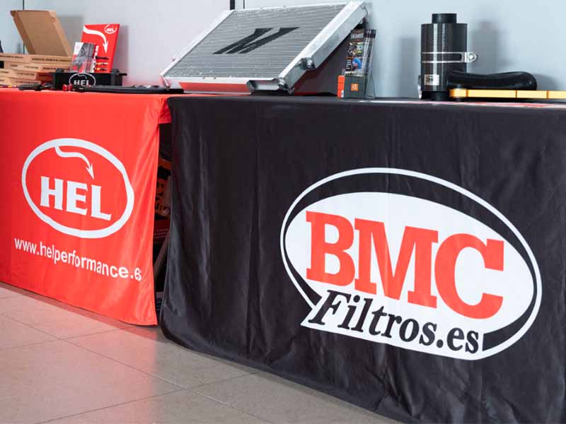 Dos mesas con pancartas de HEL y BMC Filtros.es, en las que se exponen productos y equipos de automoción para una auténtica Experiencia Profesional del Automovilismo. Encuentre las mejores soluciones de alto rendimiento en un solo lugar.