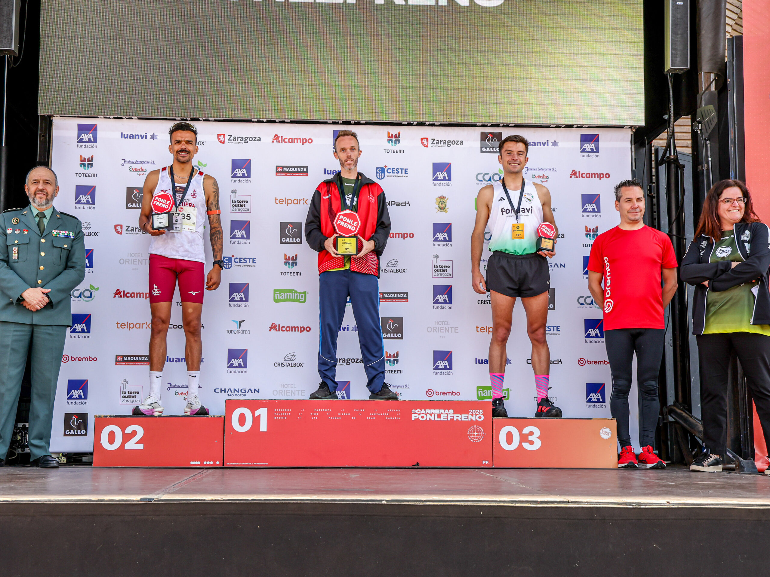 Tres atletas masculinos suben a un podio con trofeos en la mano, con personal de la prueba a ambos lados y un telón de fondo detrás.