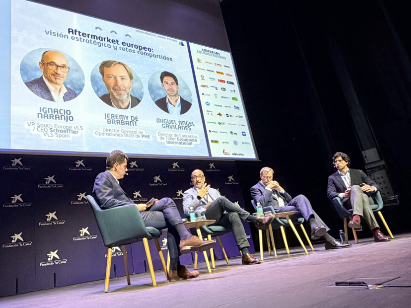 Cuatro hombres trajeados sentados en el escenario celebran una mesa redonda con una presentación proyectada detrás de ellos.