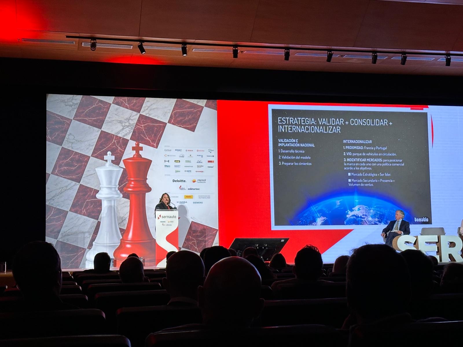 El orador hace una presentación en el escenario con un telón de fondo de temática ajedrecística y una diapositiva de estrategia; el público observa la presentación en un auditorio a oscuras.