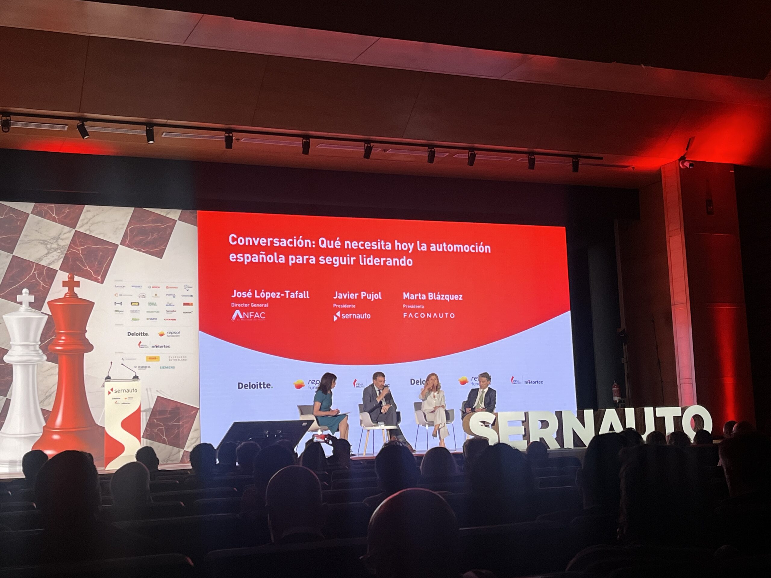 Cuatro personas se sientan en el escenario durante una mesa redonda con un telón de fondo rojo y grandes piezas de ajedrez desplegadas.