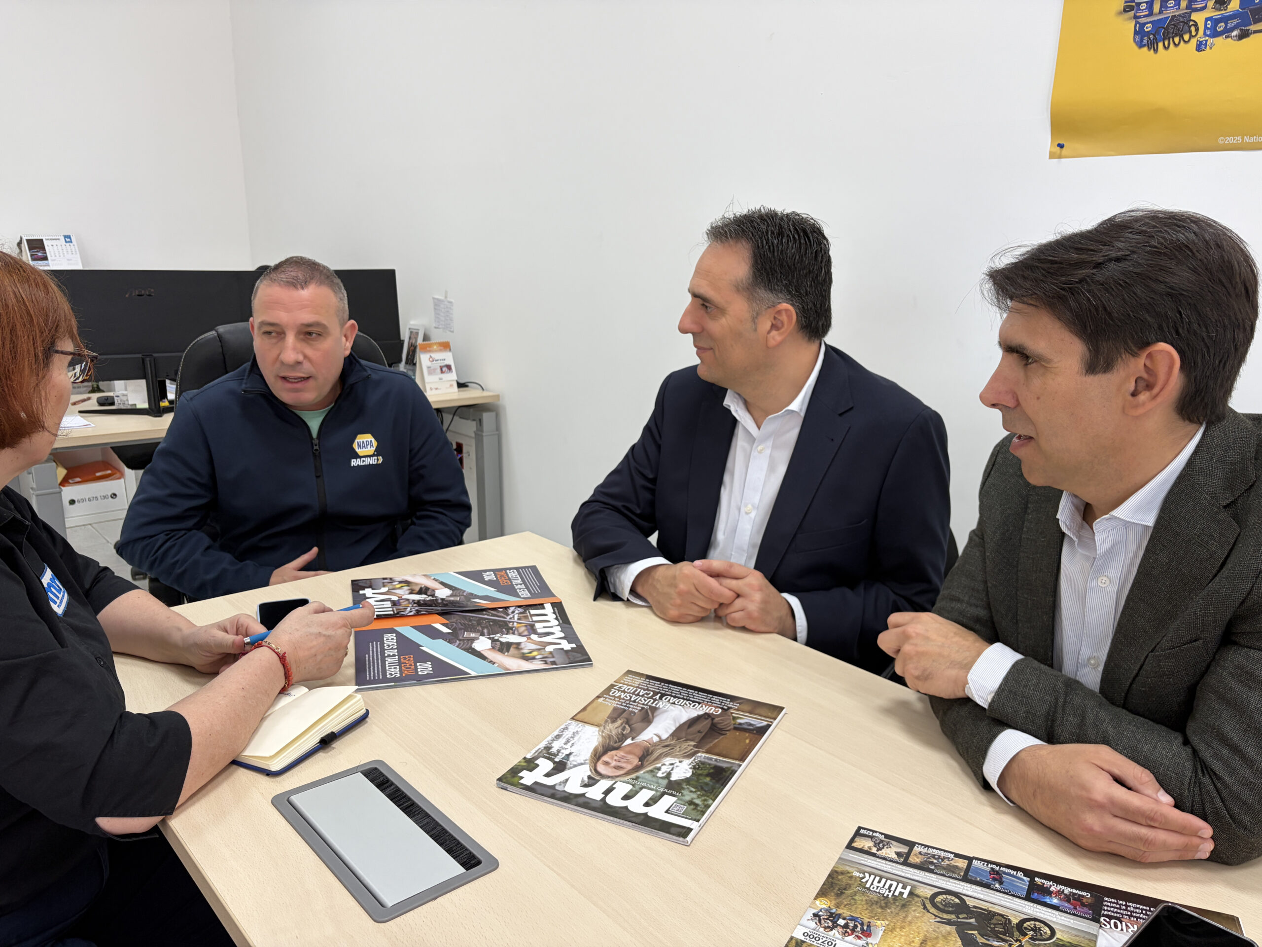Cuatro personas sentadas alrededor de una mesa manteniendo una discusión con revistas, un bloc de notas y una tableta delante.