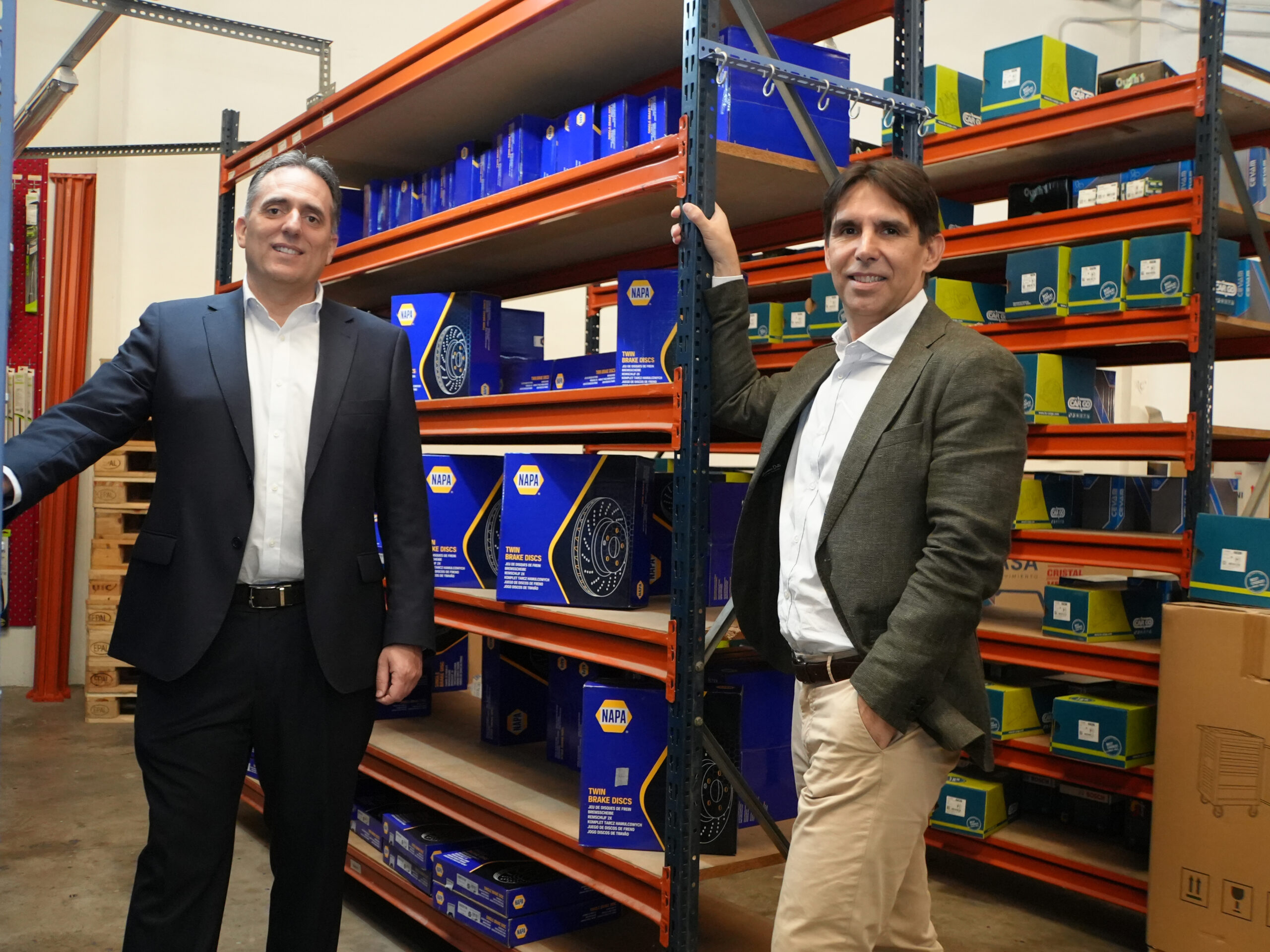 Dos hombres en traje de negocios de pie junto a los estantes abastecidos con cajas de autopartes NAPA en un almacén.