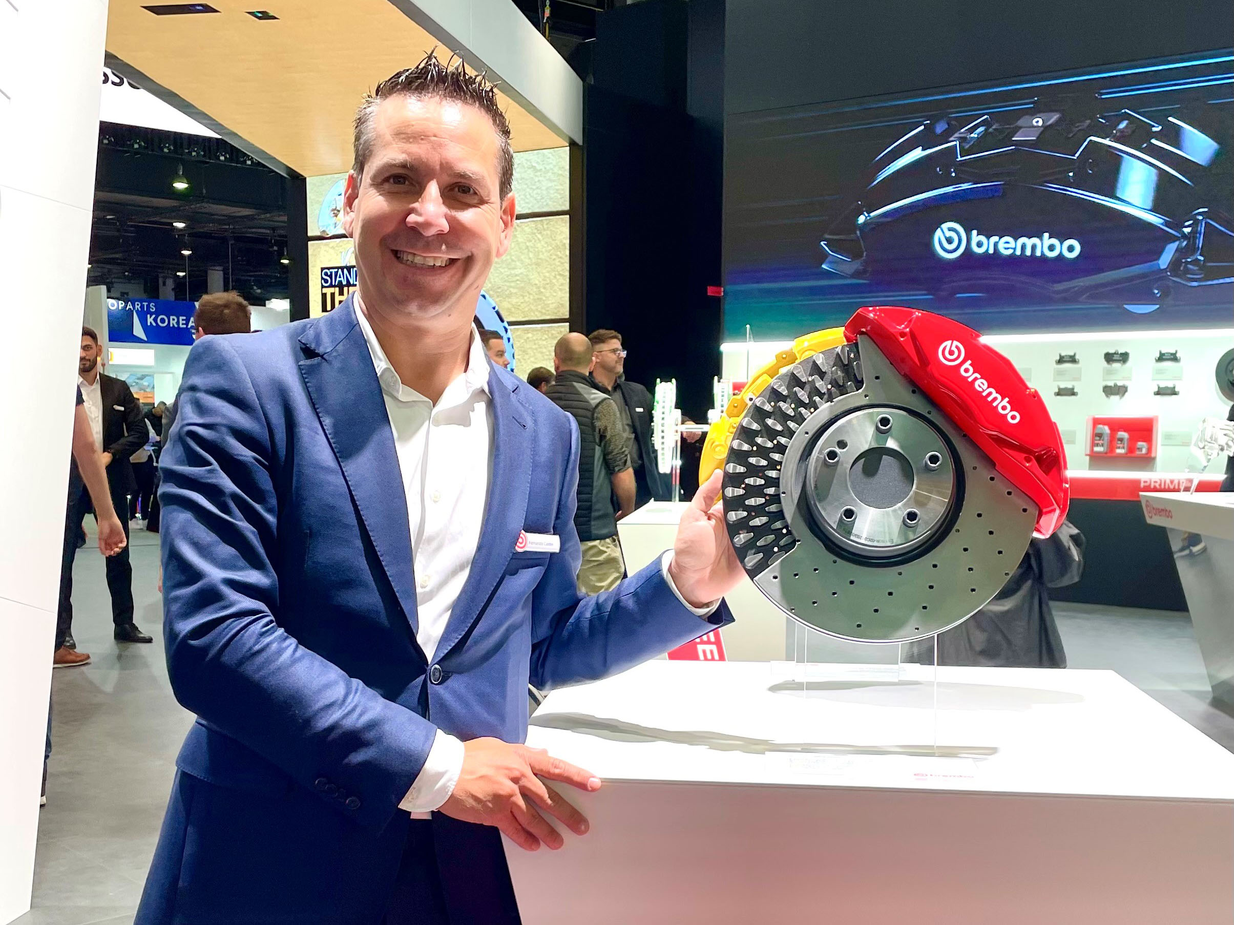 Un hombre trajeado sonríe mientras sostiene un gran disco de freno Brembo en un stand de la feria, mostrando el compromiso de la marca con la posventa de frenado y satisfaciendo la demanda sólida del mercado.