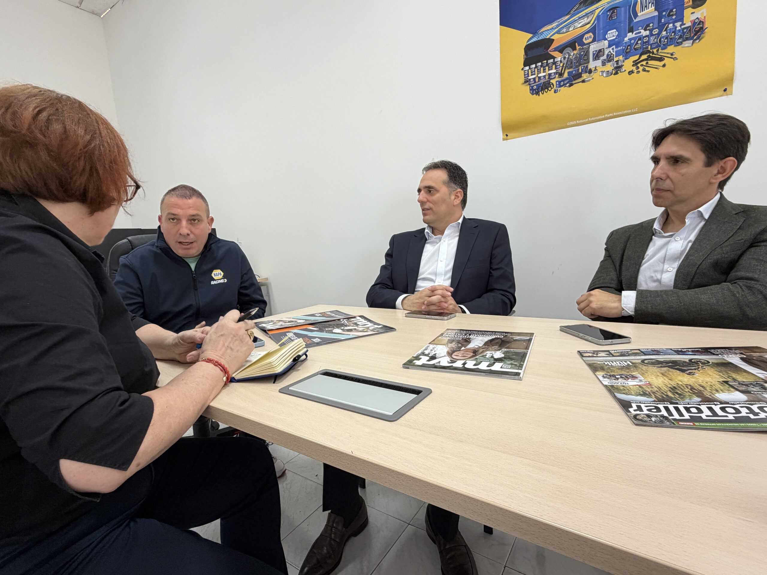 Cuatro personas se sientan alrededor de una mesa con revistas, enfrascadas en una discusión en un despacho de paredes blancas.