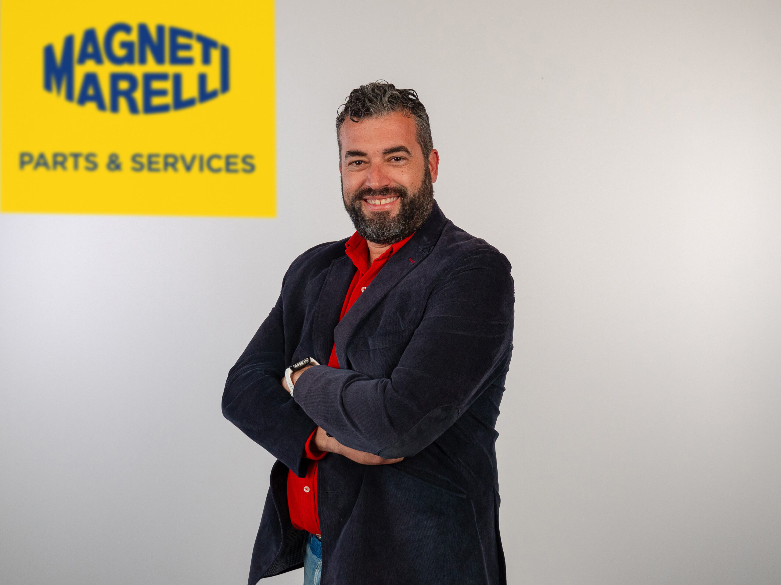 Hombre sonriente con los brazos cruzados frente a un cartel de Magneti Marelli Parts & Services en una pared blanca, destacando la demanda sólida en el sector de posventa de frenado.