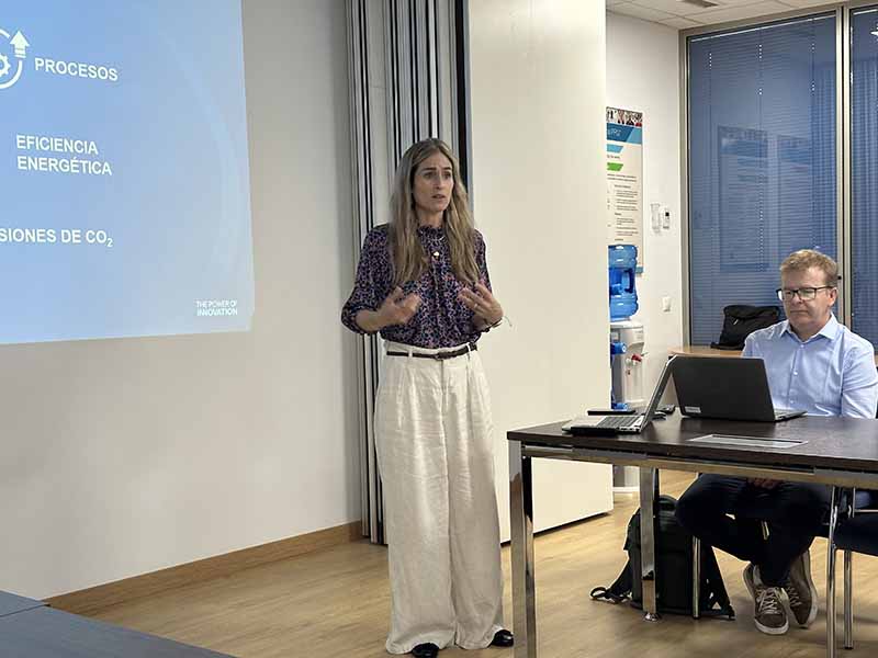 Una mujer hace una presentación junto a una pantalla de proyección, mientras un hombre está sentado en una mesa con un ordenador portátil, destacando la productividad de PPG y el eficiente sistema de mezcla Mix’n Shake en un entorno de oficina moderno.