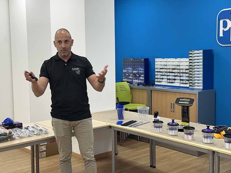 Un hombre ofrece una presentación en una sala en la que hay mesas con máscaras de seguridad, otros equipos y el barniz D8178, y destaca la productividad de PPG y el sistema Mix’n Shake.