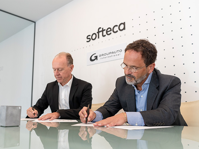 Dos hombres vestidos con traje firman unos documentos en una mesa, con los logotipos de «softeca» y «Groupauto» en la pared detrás de ellos.