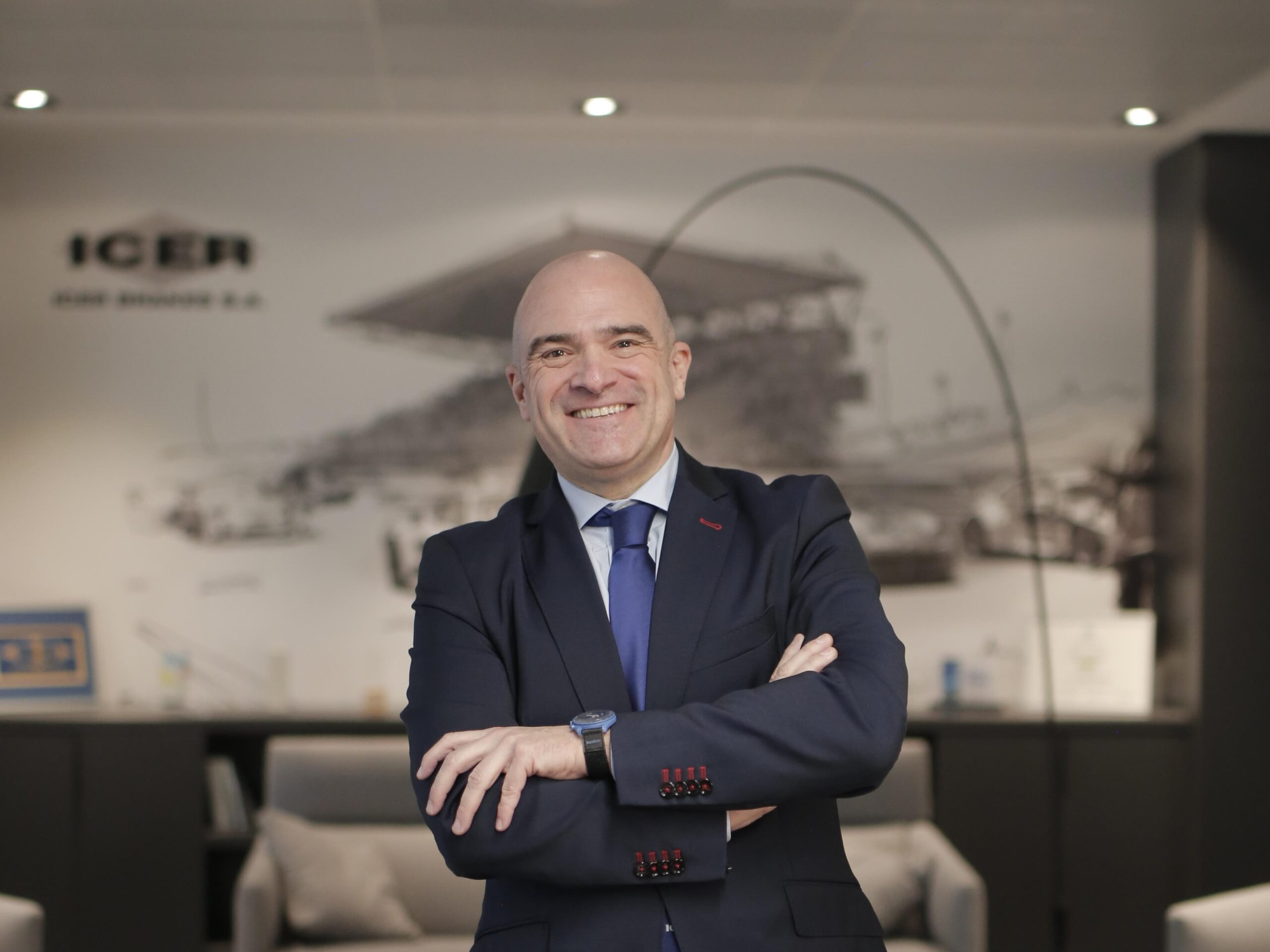Un sonriente hombre trajeado permanece de pie con los brazos cruzados en una moderna oficina con un mural de temática automovilística, reflejo de la energía del mercado y la demanda sólida en el sector posventa de frenado.
