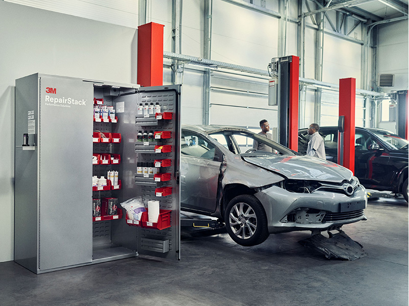 Un coche averiado en un taller de reparación, junto a un armario RepairStack de 3M abierto y repleto de material de reparación, lo que muestra cómo los talleres de carrocería pueden beneficiarse de las soluciones RepairStack Performance Solutions al mantener los productos esenciales organizados y al alcance de la mano para realizar reparaciones de forma eficiente.
