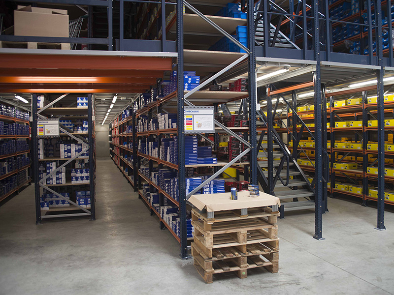 Almacén dedicado al crecimiento y la distribución, con estanterías metálicas repletas de cajas; palés y una mesita en primer plano.