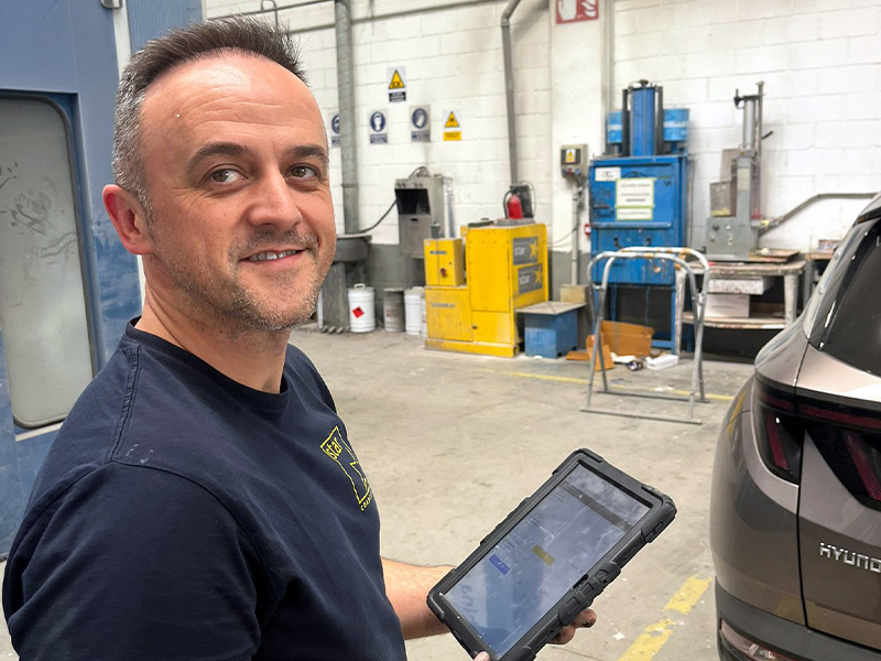 Un hombre sonriente que sostiene una tableta en un taller industrial, de pie junto a un coche, mientras utiliza iPlanner para digitalizar la actividad diaria del taller.