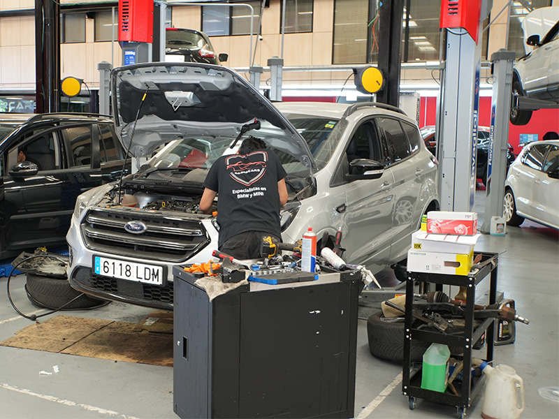 Un mecánico, reconocido entre los expertos en complejidad, trabaja en un todoterreno Ford plateado con el capó abierto en el interior de un concurrido taller de reparación de coches.
