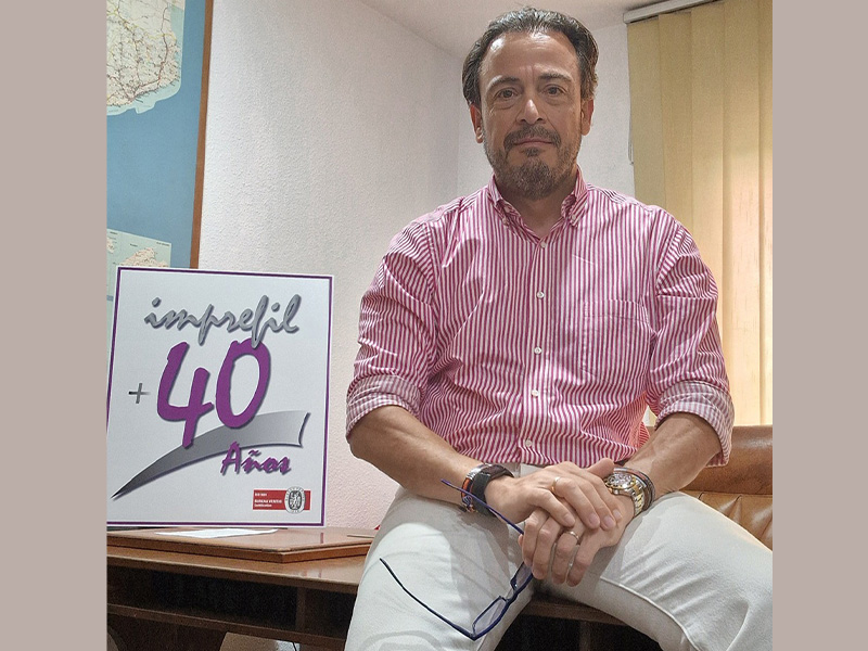 Un hombre con una camisa a rayas rojas sentado junto a un cartel en el que se lee «imprefil +40 años» en una oficina.