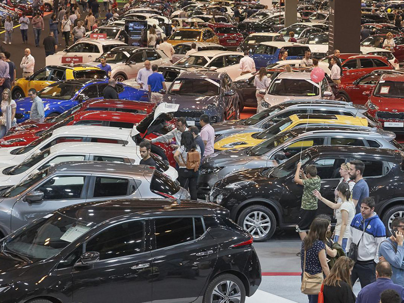 La gente pasea entre los numerosos coches aparcados en una concurrida exposición de coches cubierta o en un concesionario, admirando los últimos modelos y sus características, incluida tecnología innovadora como el borrador automático.