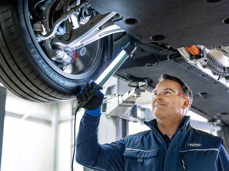 Un mecánico inspecciona la parte inferior de un coche elevado con una linterna de trabajo, con gafas de seguridad y guantes puestos, mientras se prepara para utilizar llaves neumáticas para cambiar los neumáticos en Talleres Vulco.