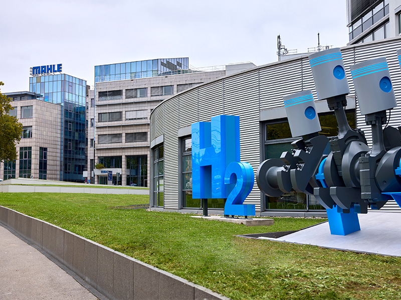 Gran escultura azul con la inscripción «H2» y una maqueta de motor en el exterior de un moderno edificio de oficinas con el letrero «MAHLE», que pone de relieve la innovadora tecnología del hidrógeno, que está cobrando impulso en toda Europa y en la Península Ibérica.