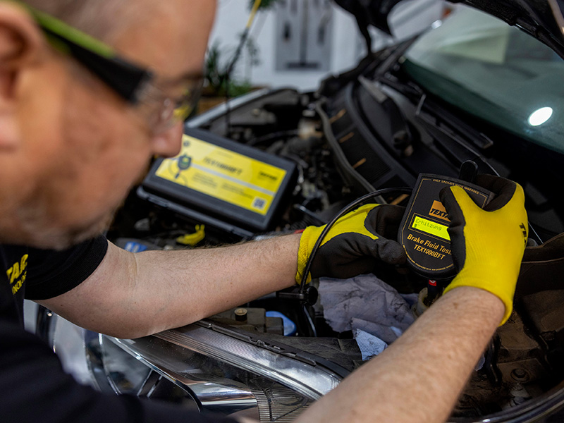 Un mecánico, que lleva guantes amarillos, comprueba el líquido de frenos de un coche con un dispositivo portátil bajo el capó, para asegurarse de que los frenos del coche funcionen de forma segura incluso bajo un calor intenso.