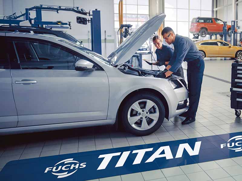 Dos mecánicos revisan el motor de un coche plateado en un luminoso taller de automóviles, perteneciente a la Red de Talleres y conocido por utilizar lubricantes FUCHS TITAN, con coches al fondo.