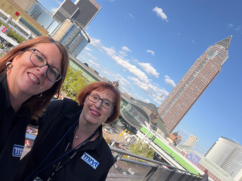 Dos mujeres con camisetas negras sonríen al aire libre, con edificios modernos y una alta torre al fondo, bajo un cielo azul, lo que refleja la expectación que genera Automechanika Frankfurt 2026, que promete más visitantes que nunca.
