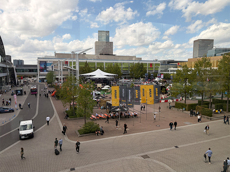 Evento al aire libre con banderas y stands; visitantes paseando entre edificios modernos con un cielo parcialmente nublado al fondo, lo que refleja el animado ambiente de Automechanika Frankfurt 2026.