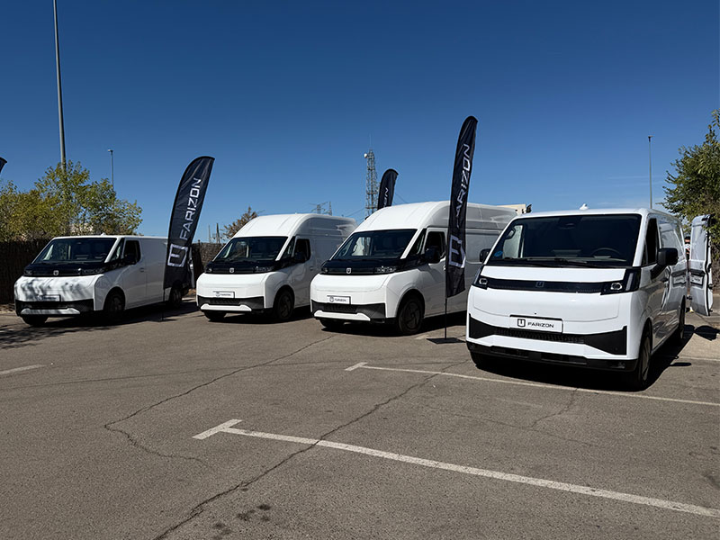 Cuatro furgonetas de reparto Farizon SV blancas, aparcadas en fila con banderas de Marizon en un día soleado en un aparcamiento, en las que se exhiben las últimas furgonetas eléctricas V7E.