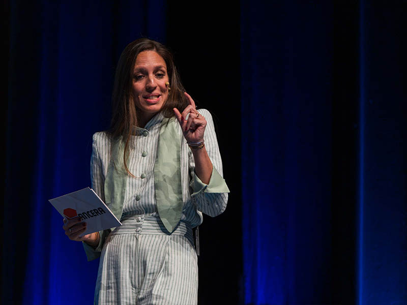 Una mujer con un traje a rayas habla en el escenario, con una tarjeta en la mano, con un telón azul como fondo.