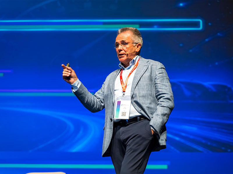 Un hombre vestido con traje habla en el escenario, con un fondo azul y futurista, y lleva una acreditación de la conferencia.