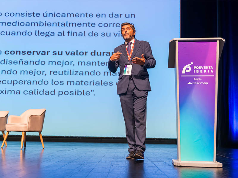 Un hombre vestido con traje habla en el escenario junto a un atril y una gran pantalla en la que se ve texto en español.