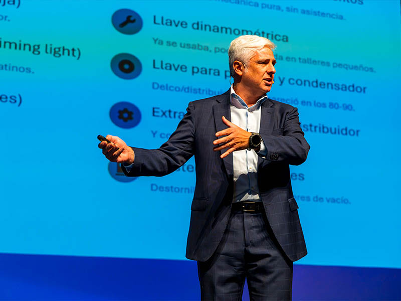 Un hombre vestido con traje dando una presentación en el escenario, gesticulando con las manos, con una pantalla azul con texto en español detrás de él.