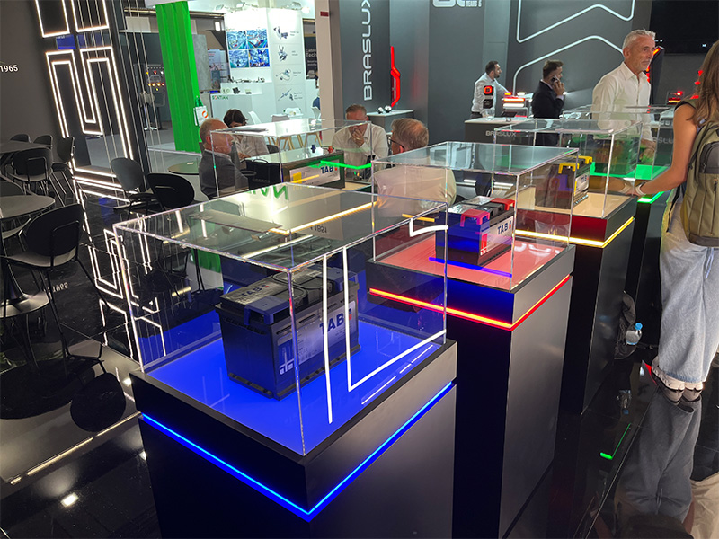 Tres baterías TAB en vitrinas de cristal con luces LED de colores, expuestas en un stand de la feria Automechanika de Fráncfort, que resaltan su nueva identidad corporativa; se ve a varias personas al fondo.