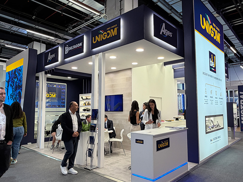 La gente se queda de pie o se sienta en un moderno stand de feria con carteles y expositores de «UNIGOM» y «Ages», donde se toman decisiones industriales que marcan el futuro del sector.