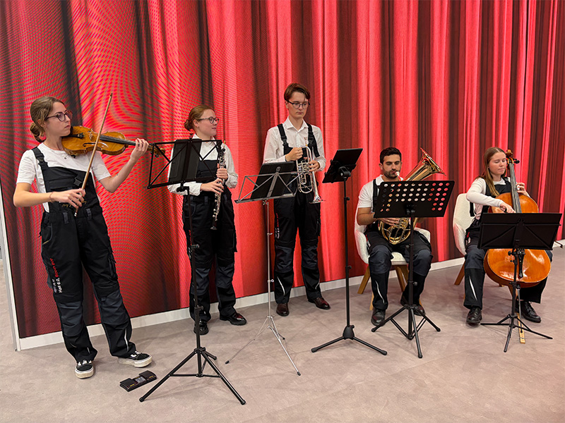 Cinco músicos tocan instrumentos de cuerda y de metal frente a un telón rojo, creando una vibrante sinfonía que recuerda a una exposición de Automechanika, presentada en colaboración con febi.