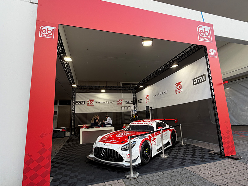 Coche de carreras rojo y blanco expuesto en un stand rojo con la marca febi y pancartas, en el interior, sobre un suelo con diseño de bandera a cuadros.