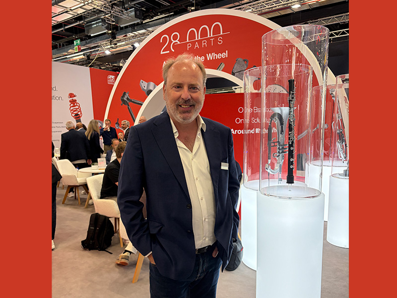 Un hombre con una chaqueta azul marino posa sonriente en un stand de la feria Automechanika, con los recambios y expositores de febi como telón de fondo, creando una sinfonía de innovación y excelencia automovilística.