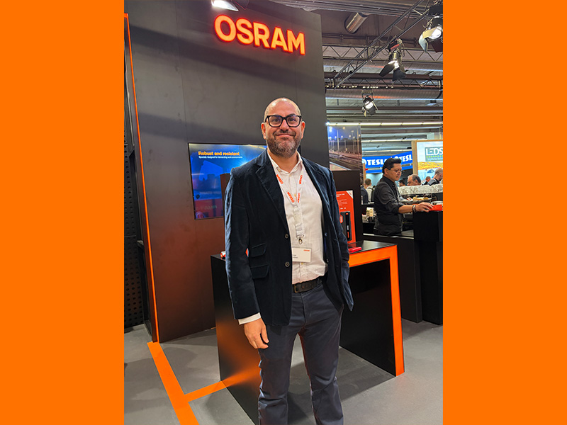 Un hombre con chaqueta de traje está junto a un expositor de Osram en el que se muestra un borrador eléctrico en una feria comercial.